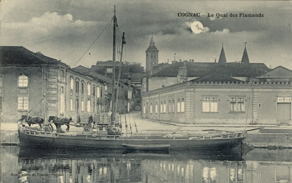 Old postcard of the Quai des Flamands on the banks of the Charente in Cognac, with a gabare (flat-bottomed boat) in the foreground, carrying cases of bottles of Cognac, and the old town and the bell tower of Saint Léger church in the background