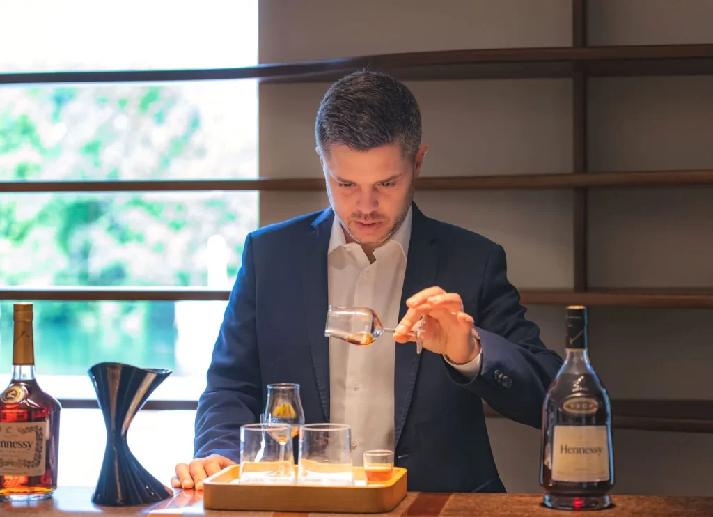 Homme tenant délicatement dans sa main un verre de cognac à l'horizontal en observant le cognac, de chaque côté une bouteille de cognac de la marque Hennessy au circuit de visite à Cognac